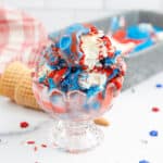 A glass bowl of red, white and blue ice cream with cones and sprinkles around it.