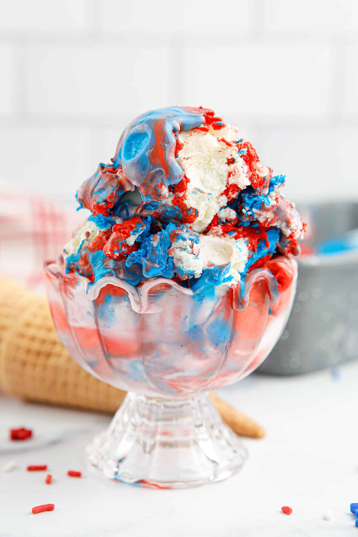 A glass bowl of red, white and blue ice cream with cones and sprinkles around it.