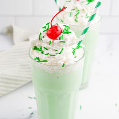 Two green Shamrock Shakes topped with whipped cream, cherries and sprinkles on a white background with a striped towel in the background.