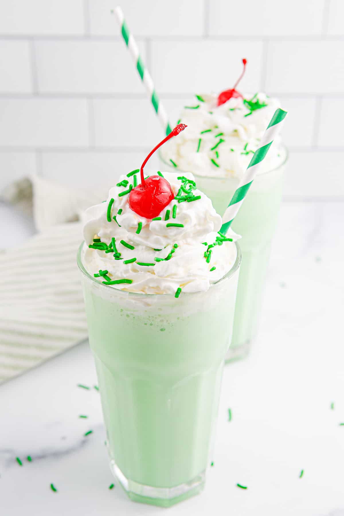 Two milkshake glassed filled with green shamrock shake topped with whipped cream, cherries and green sprinkles with green and white straws in them and a light green and white towel in the background.
