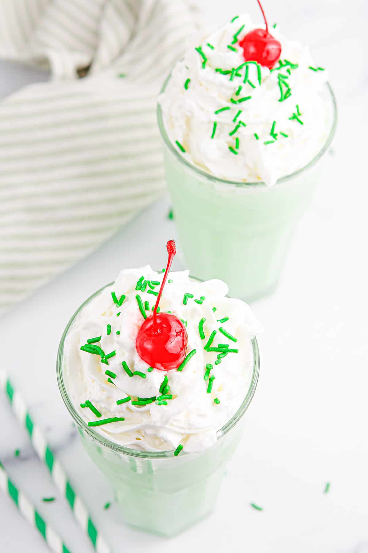 Two green shamrock shakes topped with whipped cream, cherries and sprinkles on a white background with a light green and white striped towel.