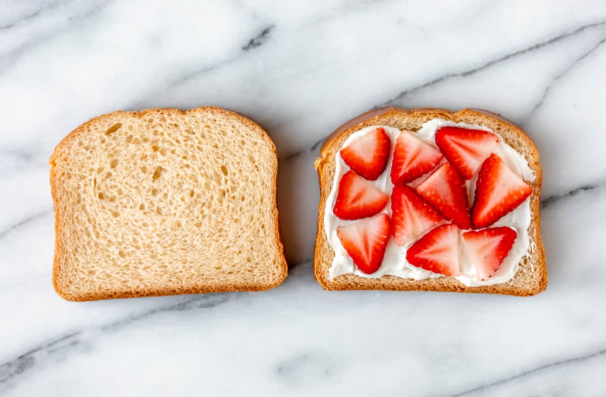 Two slices of brioche bread with cream cheese and slices of strawberries on one of the slices over a marble background.