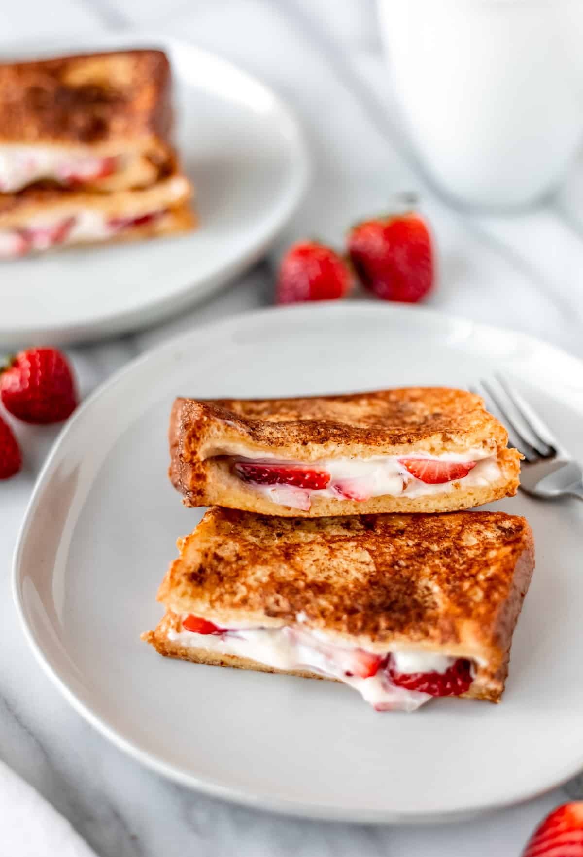 Two halves of strawberry french toast layered over one another on a white plate with a mug, strawberries and a second plate of french toast in the background.