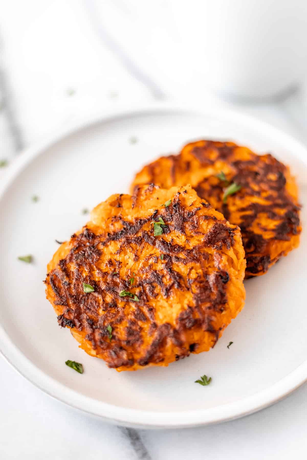 Two sweet potato hash browns layered on a white plate with a white mug in the background.
