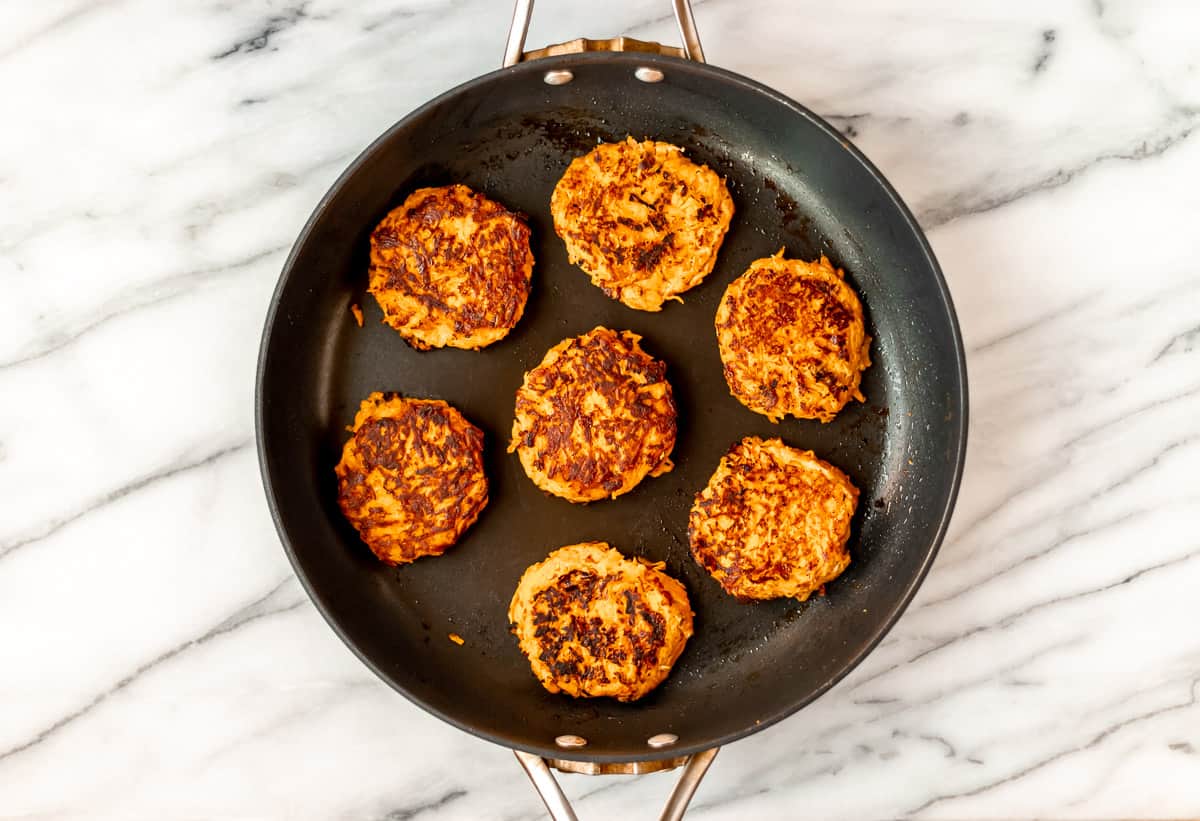 Seven sweet potato hash browns cooking in a black skillet.