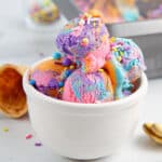 A white bowl filled with scoops of Unicorn Ice Cram with sprinkles and cones around it.