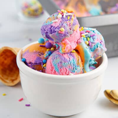 A white bowl filled with scoops of Unicorn Ice Cram with sprinkles and cones around it.