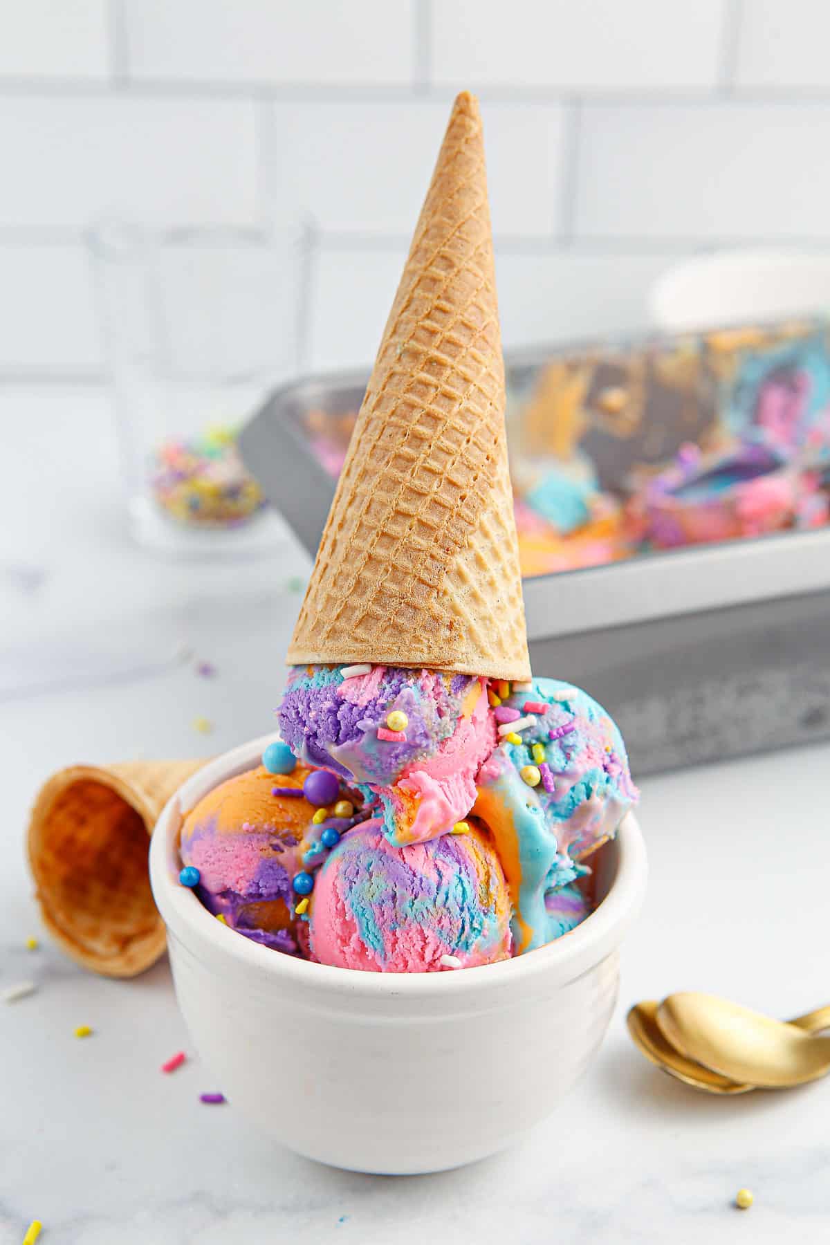 A bowl of multi-colored unicorn ice cream in a white bowl topped with an ice cream cone.