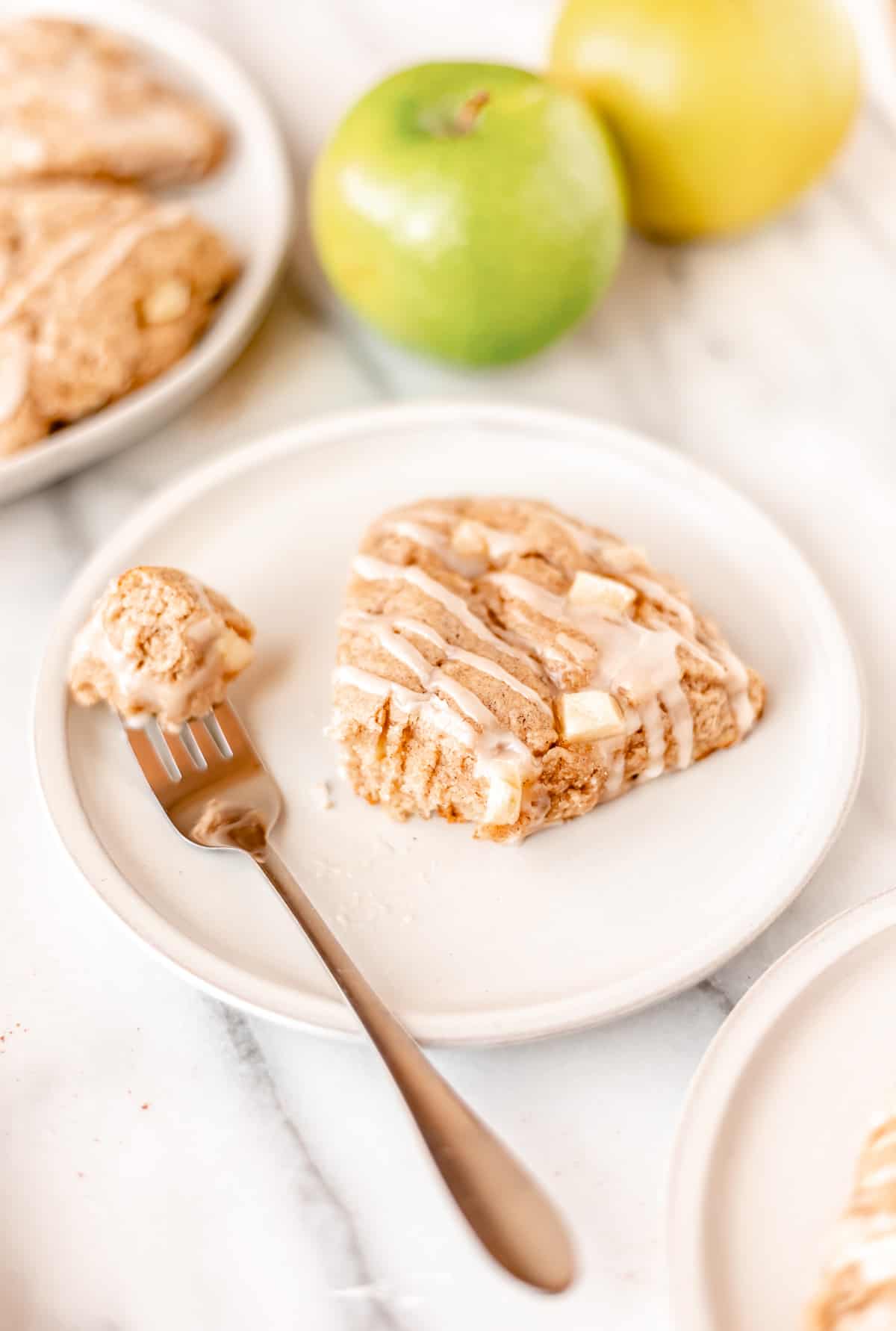 An apple cinnamon scone on a white plate with a bite taken out on a fork next to it with a second plate and apple in the background.