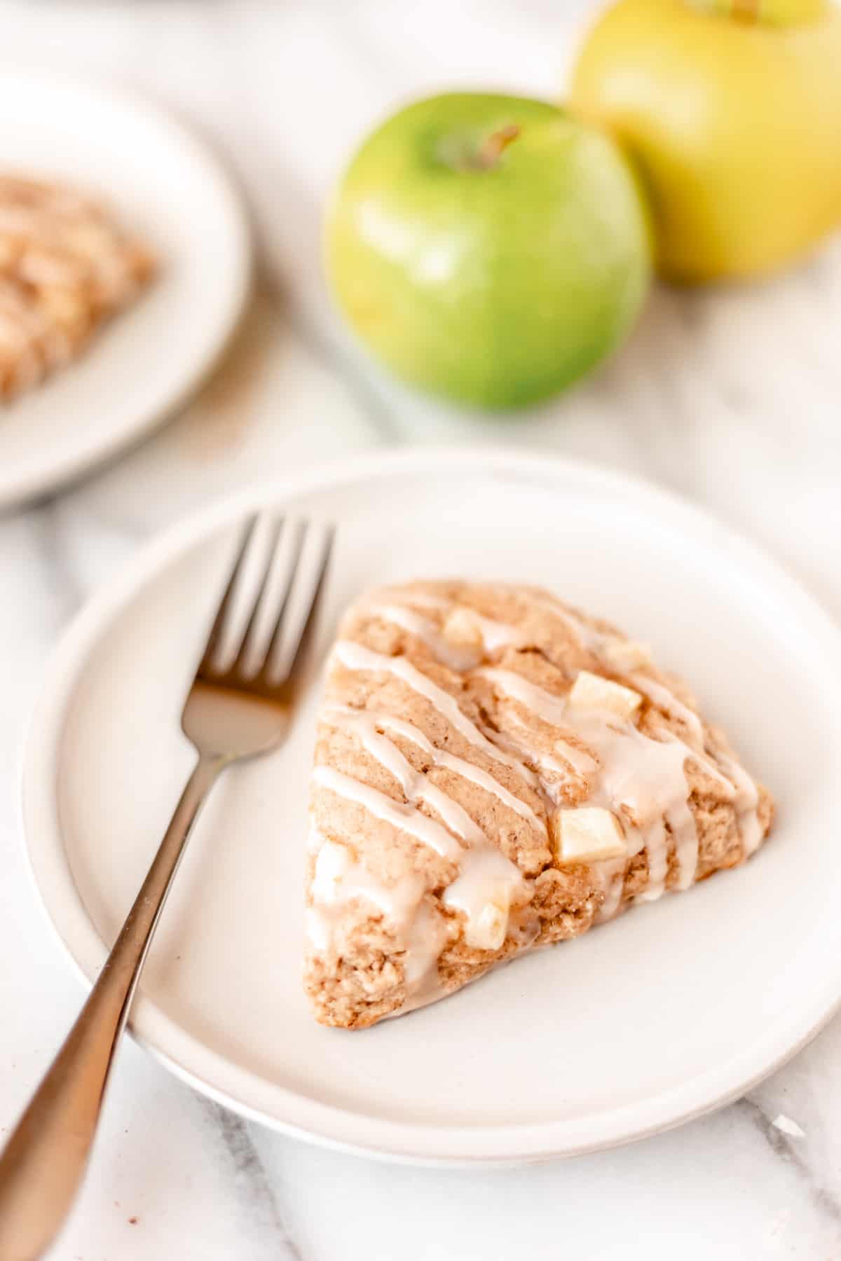 An apple cinnamon scone on a white plate with a fork next to it with a second plate and apples in the background.