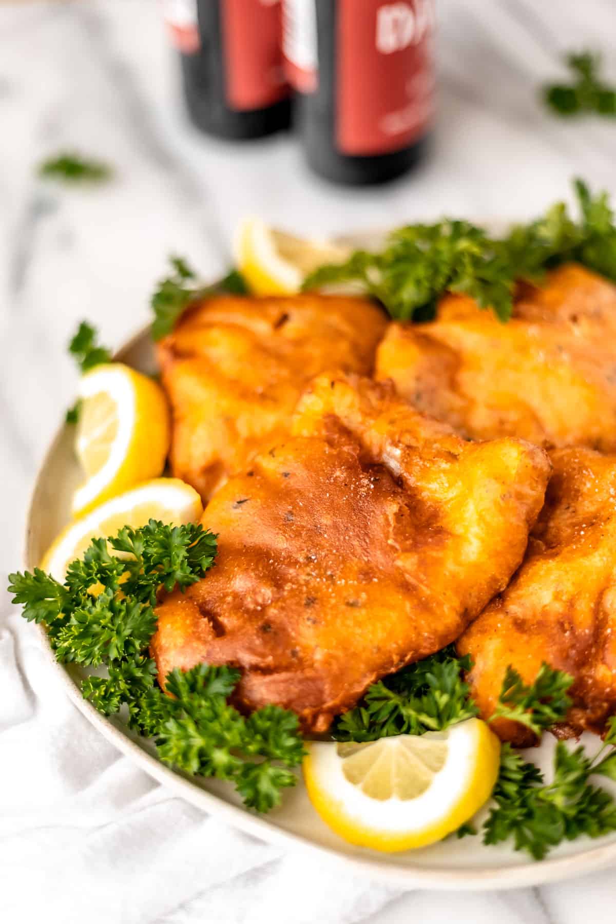 Close up of beer battered cod fillets on a plate with parsley and lemon slices with two bottles of beer and more parsley in the background.