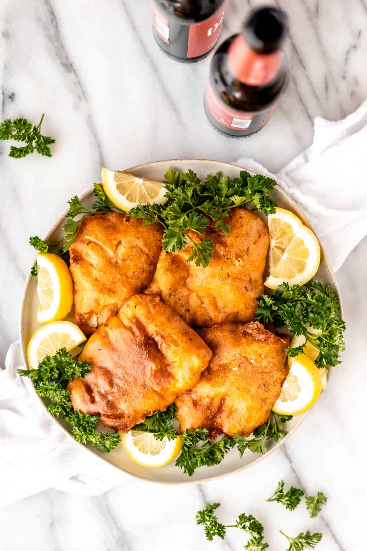 Overhead of beer battered cod fish on a plate with lemons and parsley with two bottles of beer and parsley around it.