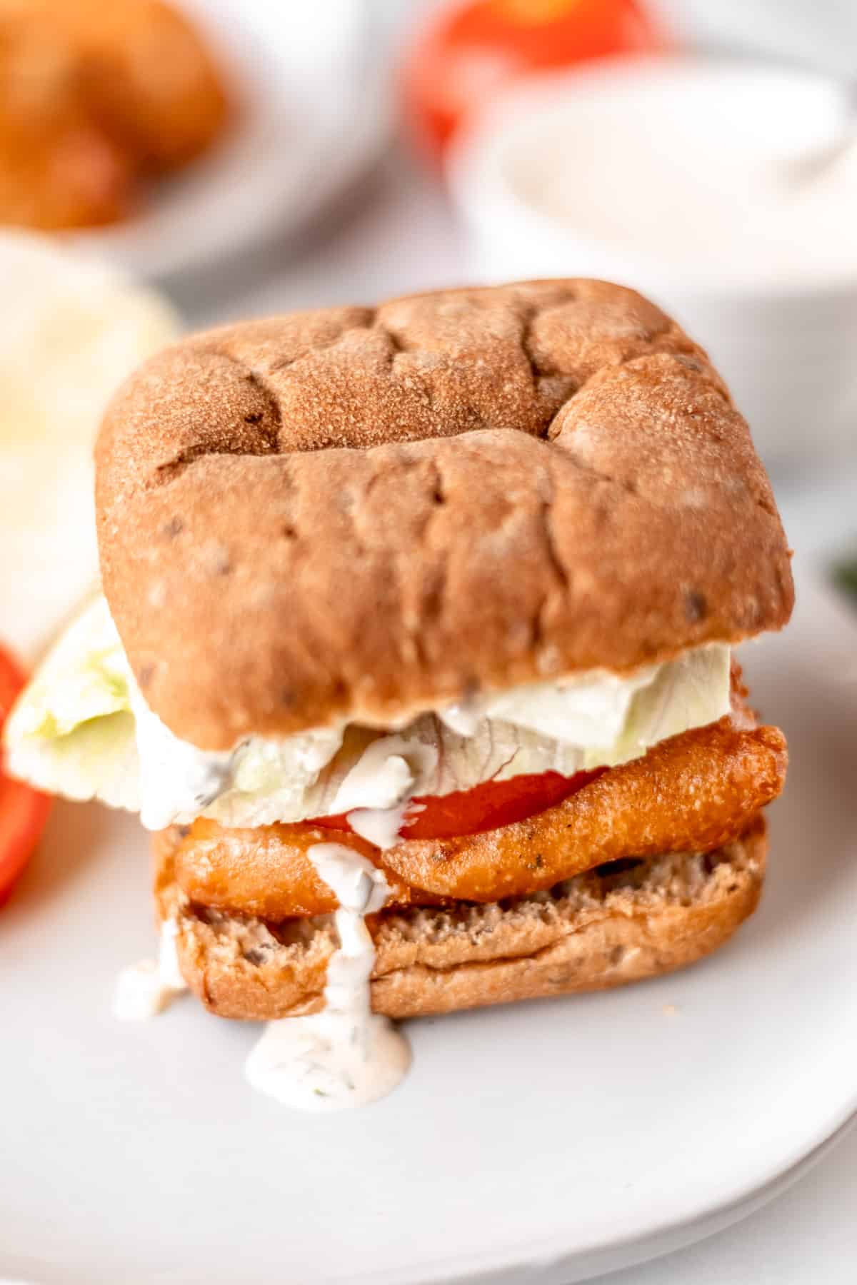A cod sandwich with fried fish, lettuce, tomato and tartar sauce on a white plate with ingredients blurred in the background.