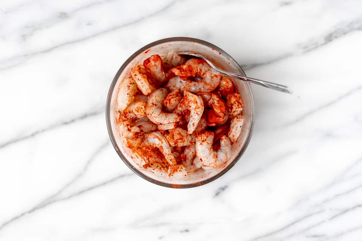 Seasoned shrimp with paprika in a glass bowl with a spoon in it on a marble background.