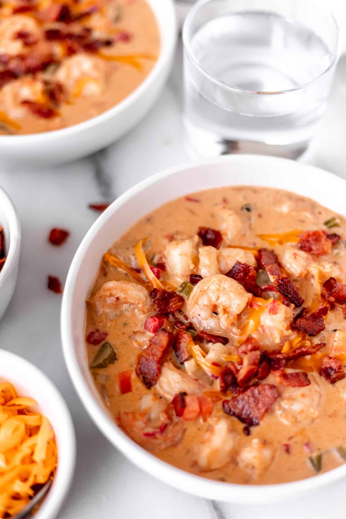 Two bowls of creamy shrimp chowder in white bowls with a glass of water, bowl of cheese and crumbled bacon around them.