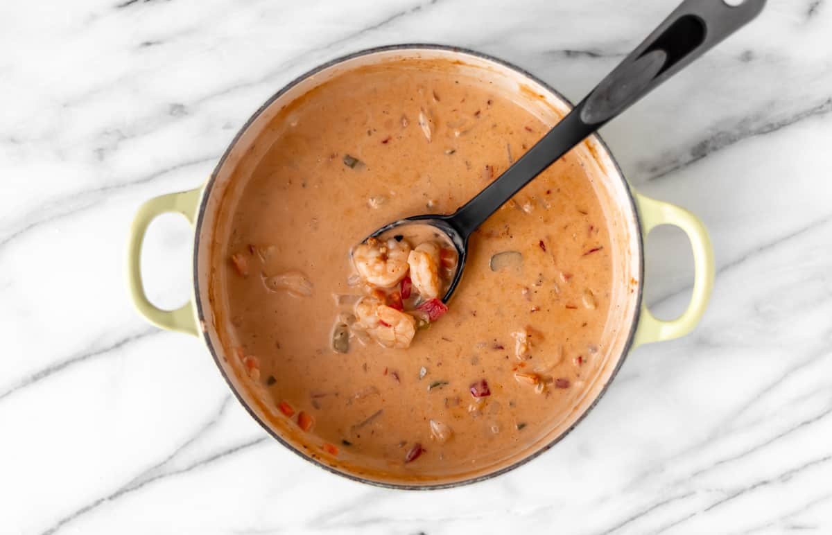 Creamy shrimp soup in a Dutch oven with a spoon lifting up some of the shrimp and vegetables over a marble background.