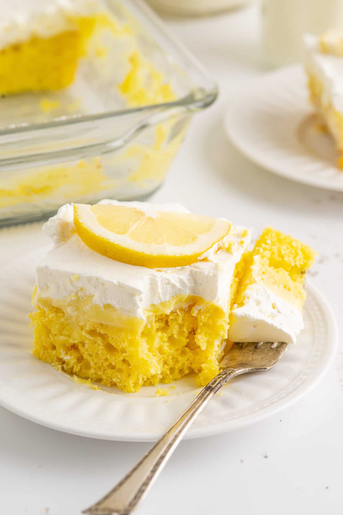 A serving of lemon poke cake on a white plate with a fork with a bite on it.