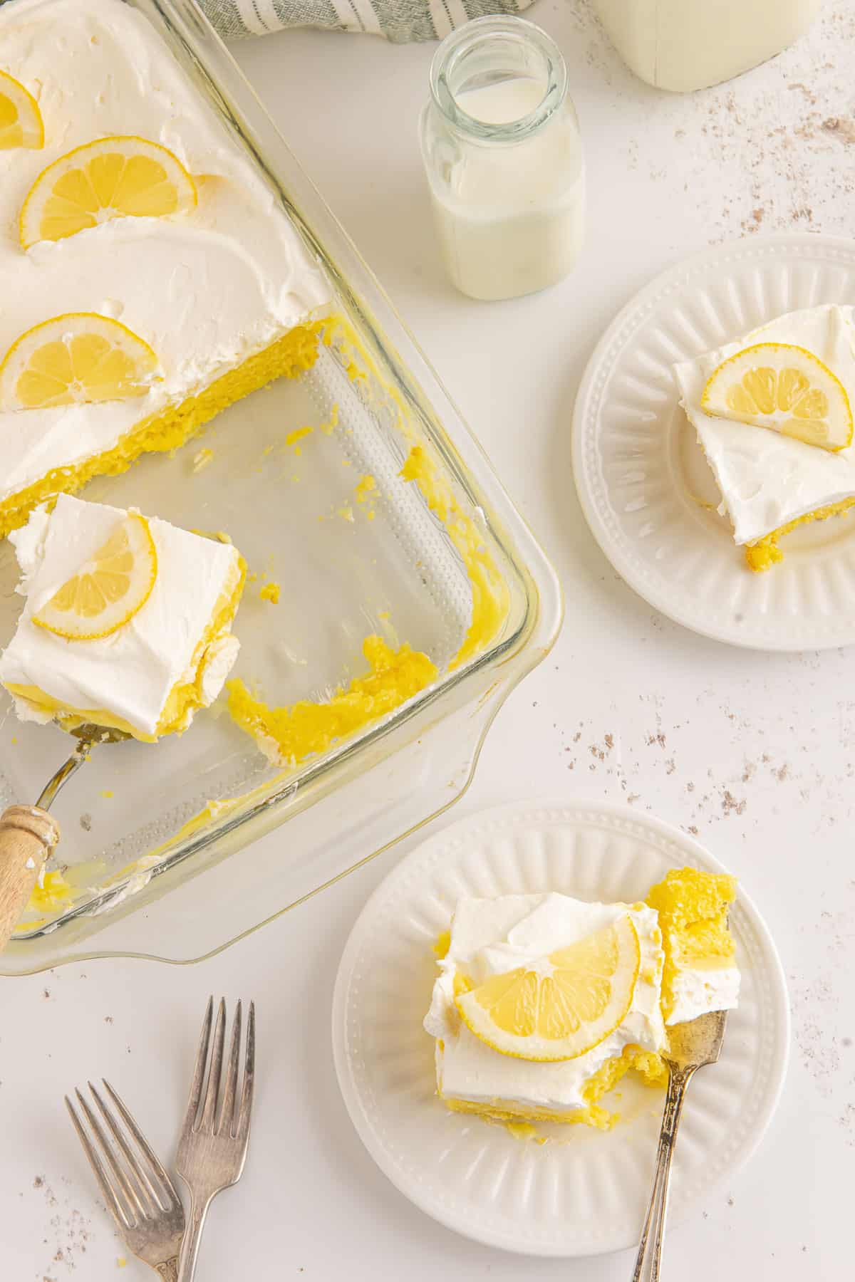 Overhead of multiple servings of lemon poke cake on plates and in the baking dish with forks, milk and a towel around them.