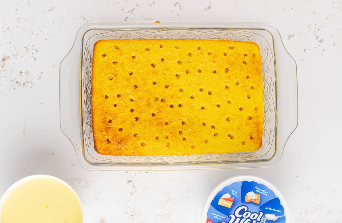 Baked lemon cake with holes poked into it with a bowl of pudding and a tub of cool whip around it.