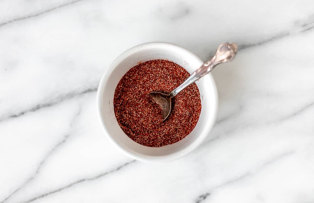 Spices mixed together in a small white bowl with a spoon on a marble background.