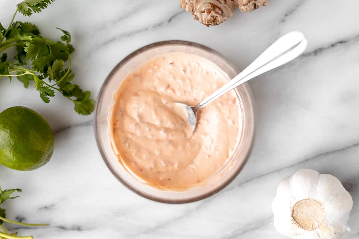 Spicy mayonnaise in a glass bowl with a spoon in it over a marble background with other ingredients around it.