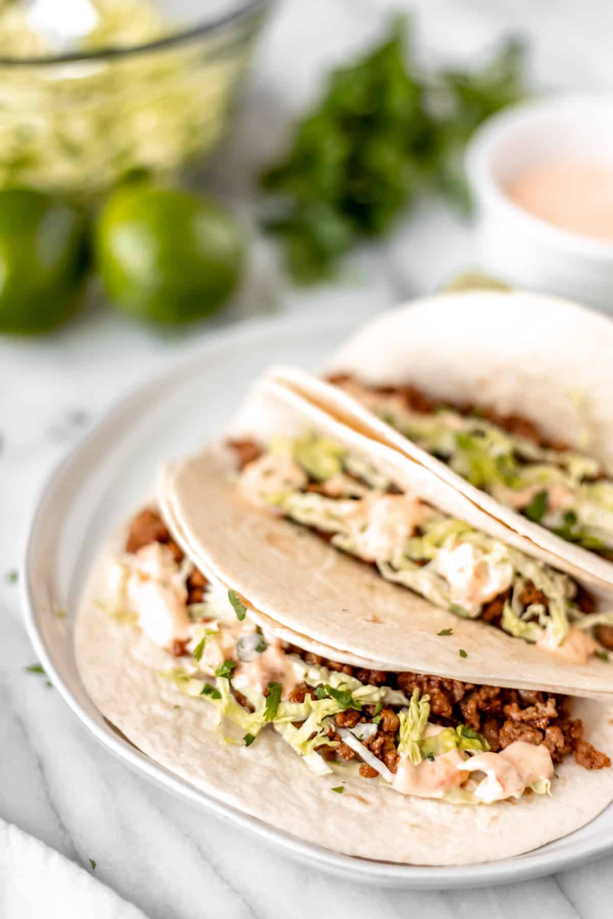 Three Asian pork tacos on a white plate with cilantro and lime in the background,