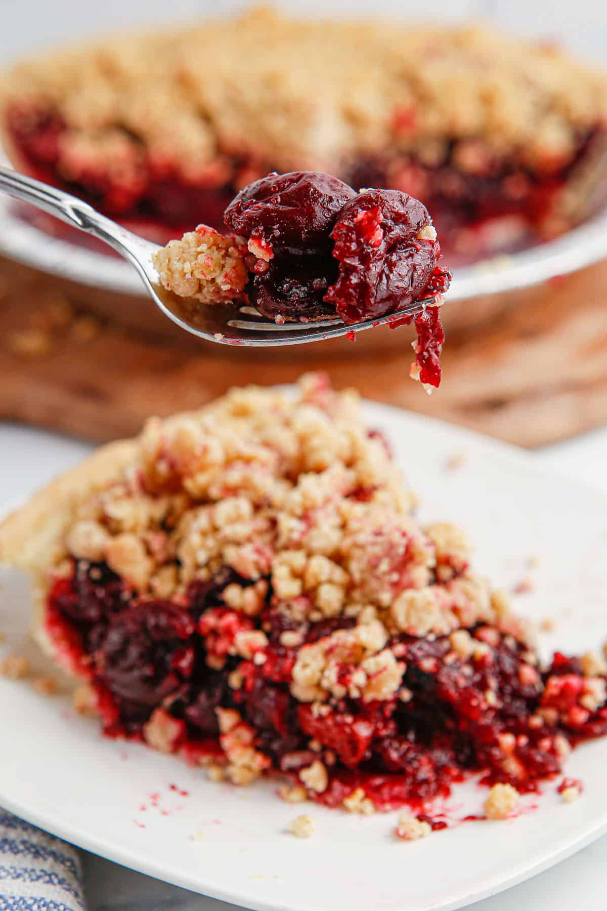 A slice of cherry crumb pie on a white plate with a fork lifting up a bite.