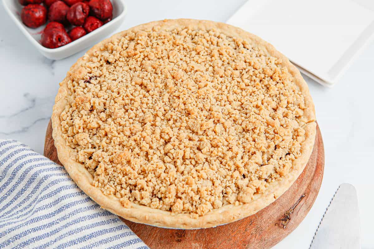 A baked cherry crumb pie on a wood board with a bowl of cherries and striped towel around it.