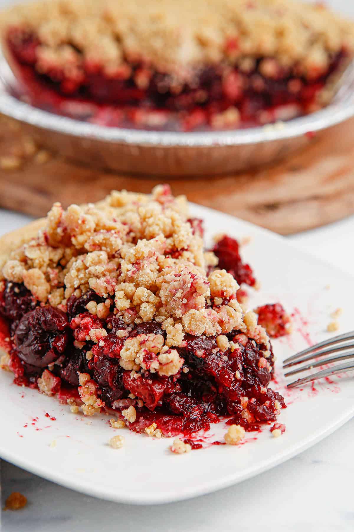 A slice of cherry crumb pie on a white plate with a fork with the remainder of the pie in the background.