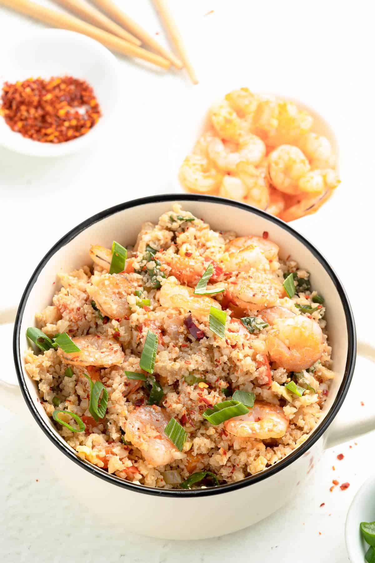 A bowl of shrimp cauliflower fried rice with a bowl of shrimp and a bowl of seasoning in the background.