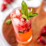 A strawberry mojito in a glass with fresh mint on a wood server with strawberries around it.