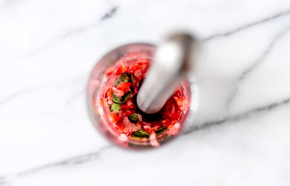 Strawberries and mint in the bottom of a glass with a muddler in it over a marble background.