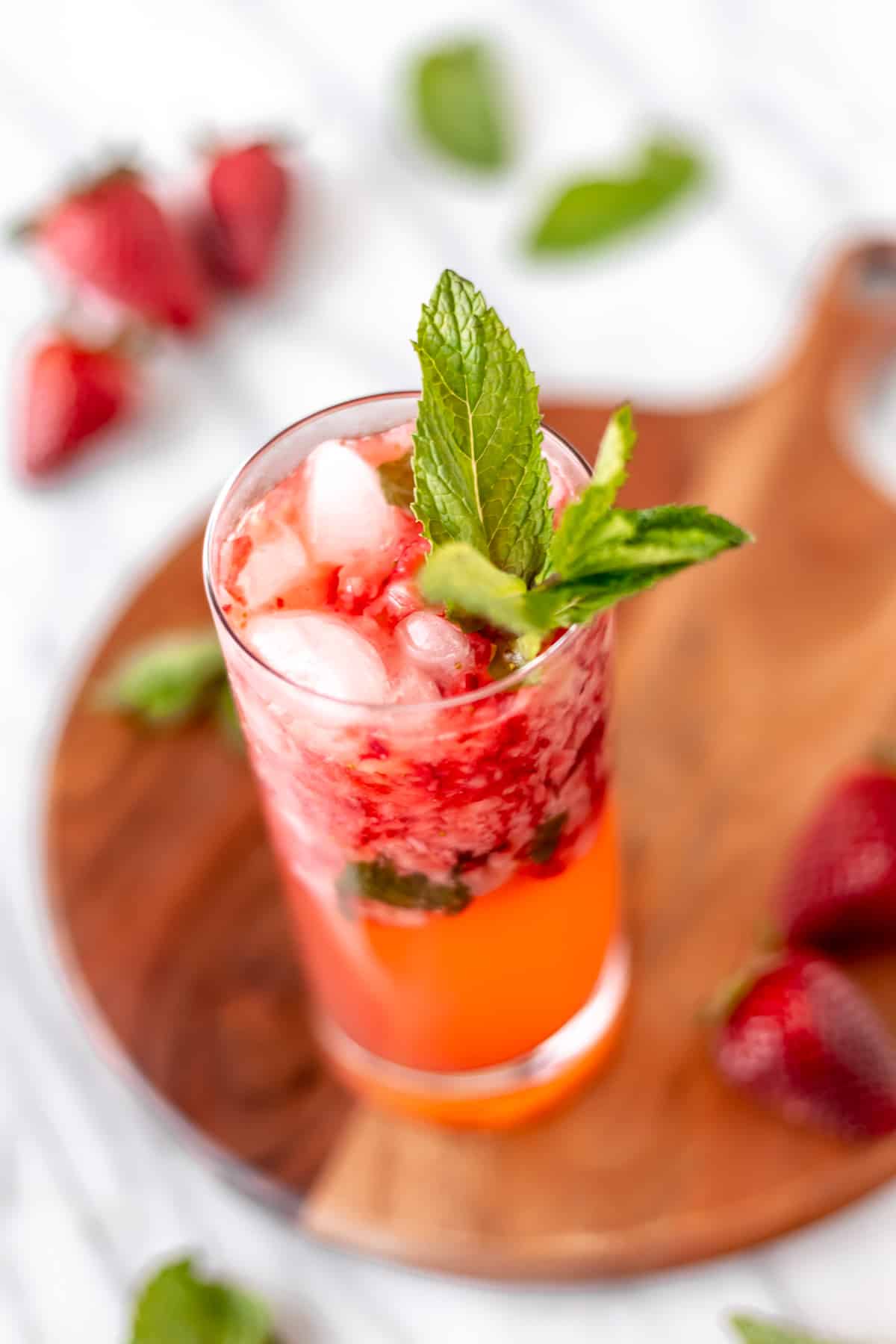 A strawberry mojito in a glass with fresh mint on a wood server with strawberries and mint leaves around it.