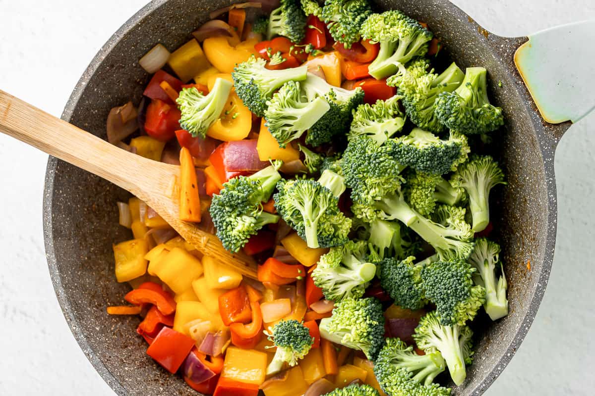 Peppers, onion and broccoli in a skillet with a wood spoon.