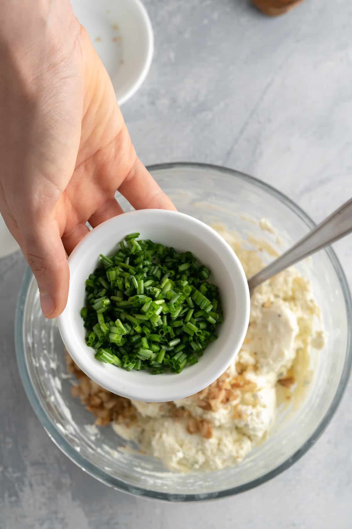 Cream cheese mixture in a bowl with a hand holding up a small bowl of chives to be mixed into it.