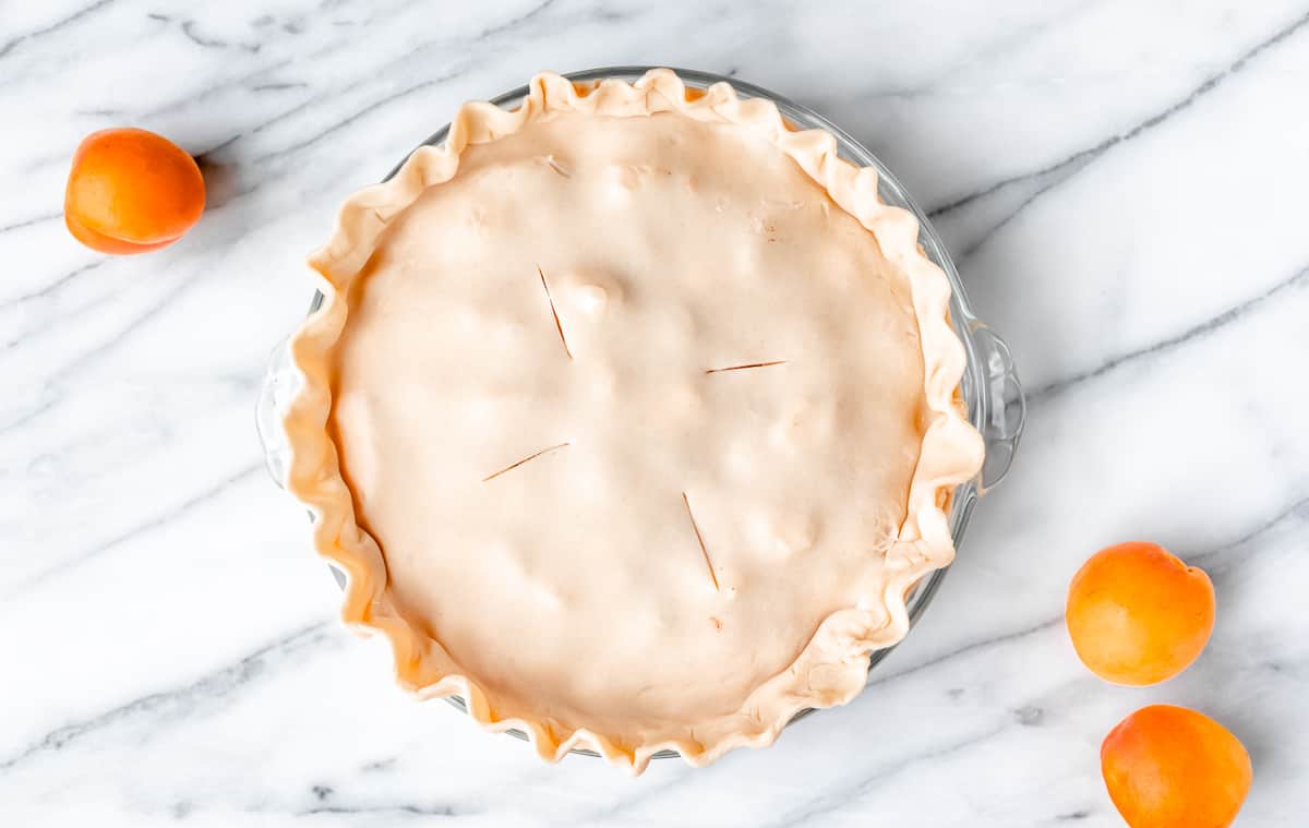 An unbaked apricot pie on a marble background with three apricots around it.