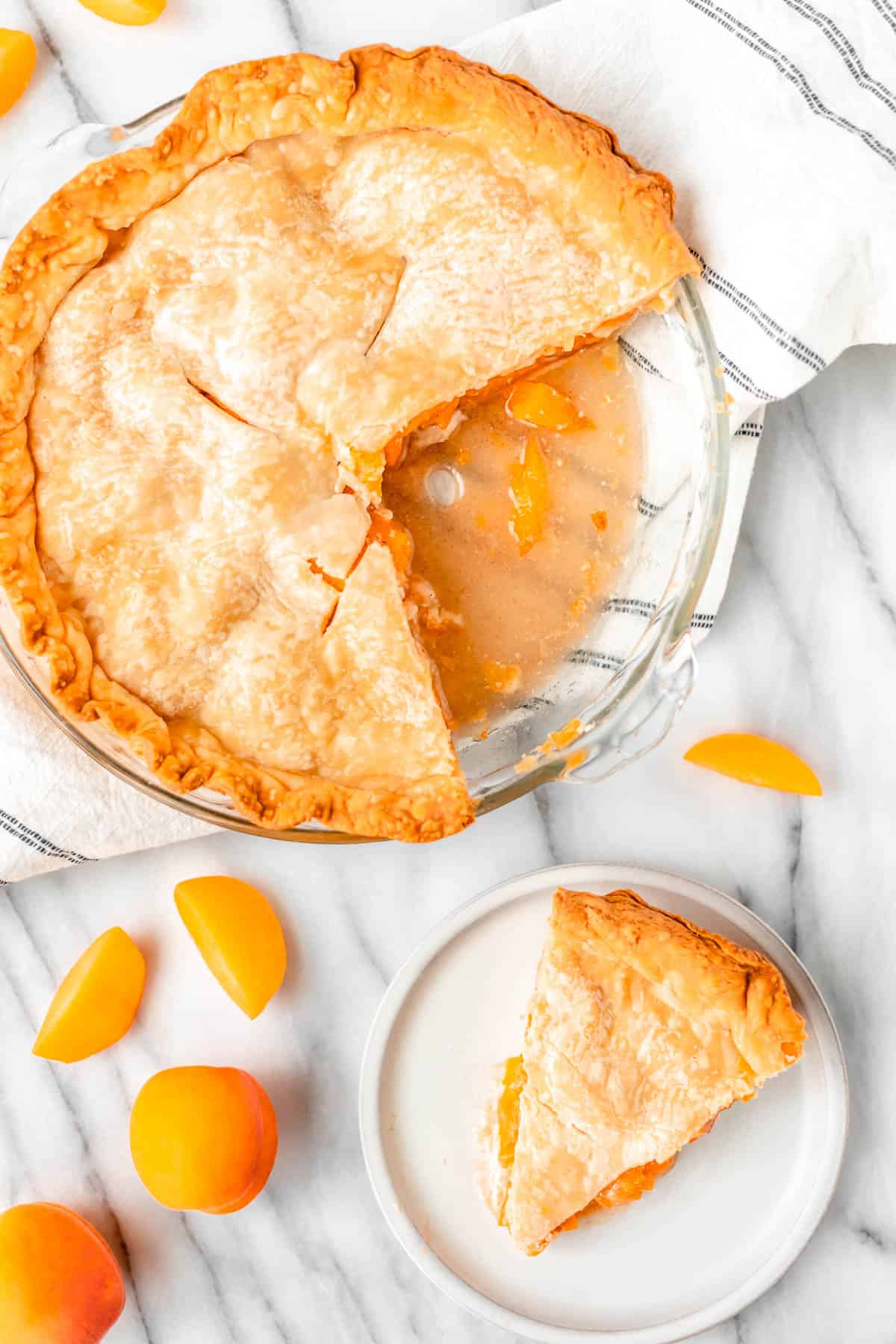 Overhead of an apricot pie with a slice on a white plate and apricots around it.