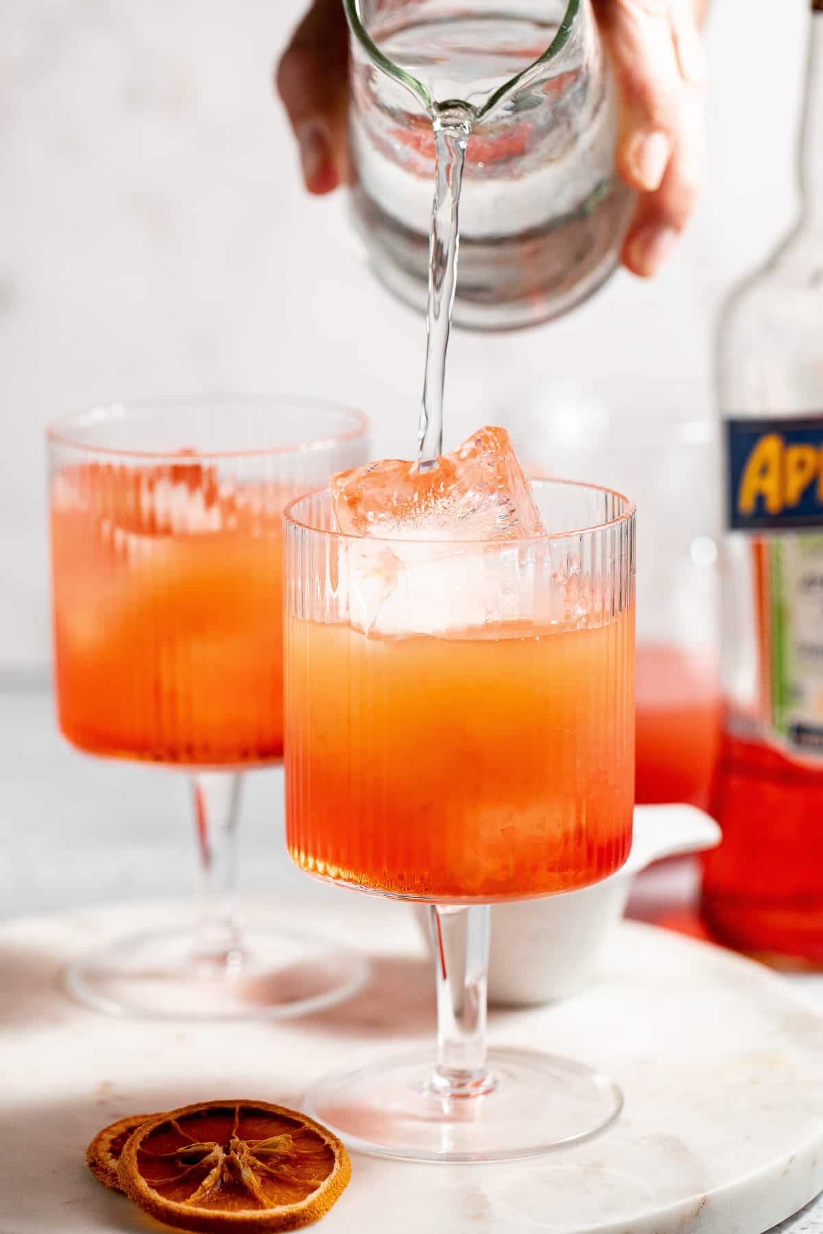 Club soda being poured over an aperol spritz with a second drink and aperol bottle in the background.