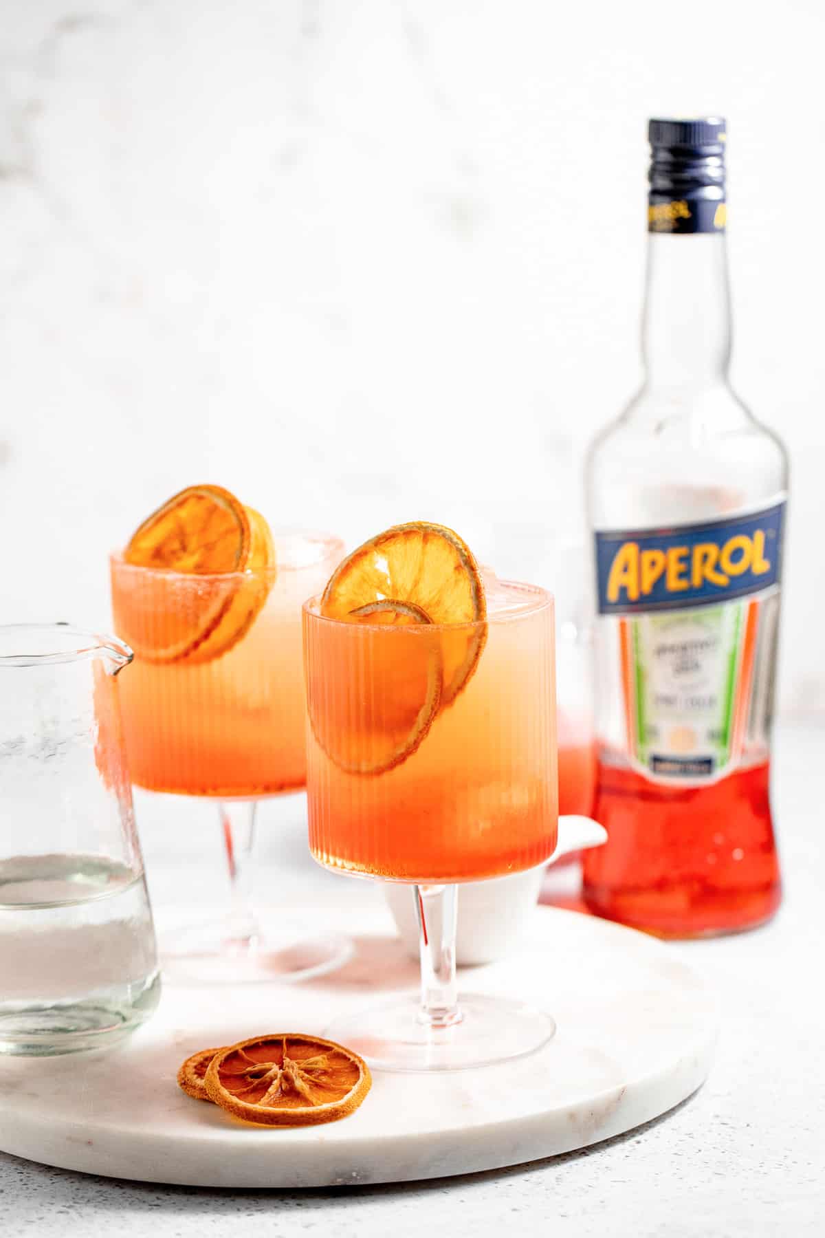 2 glasses of blood orange aperol spritz's on a white tray with a bottle of Aperol in the background.