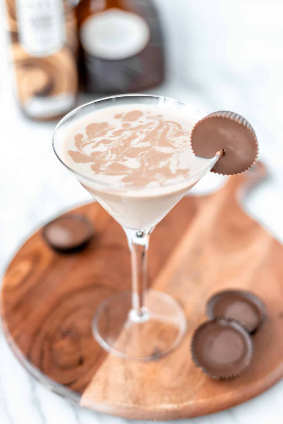 A chocolate peanut butter martini on a wood server with Reese's cups around it and bottles blurred in the background.