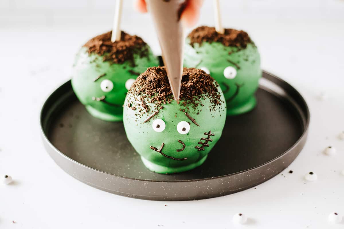 Three Frankenstein candy apples on a black plate with a piping bag close to one.