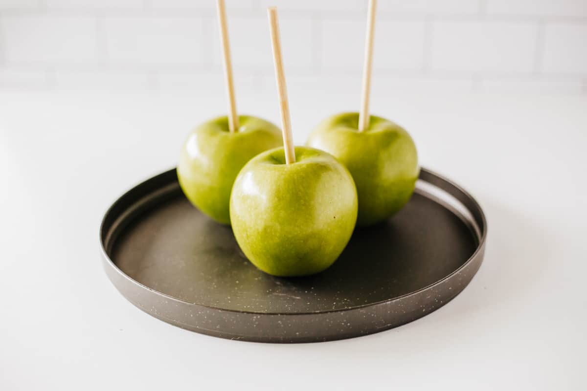 Three green apples on a plate plate with bamboo sticks in the tops of them.