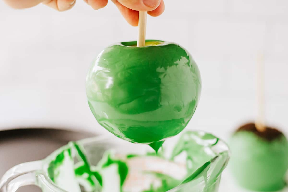 An apple that's been dipped in green candy melts being held up over a measuring cup.