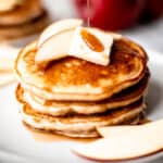A stack of apple cinnamon pancakes with syrup being poured onto with apples and a second plate in the background.