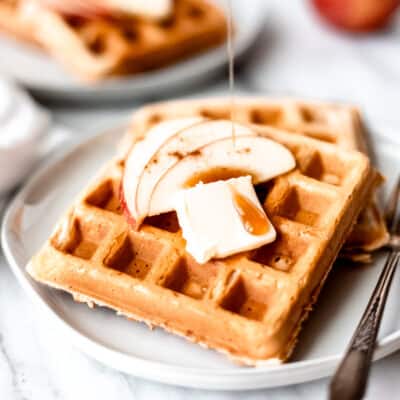 Apple cinnamon waffles on a plate with maple syrup being poured over them and a second plate and apple in the background.