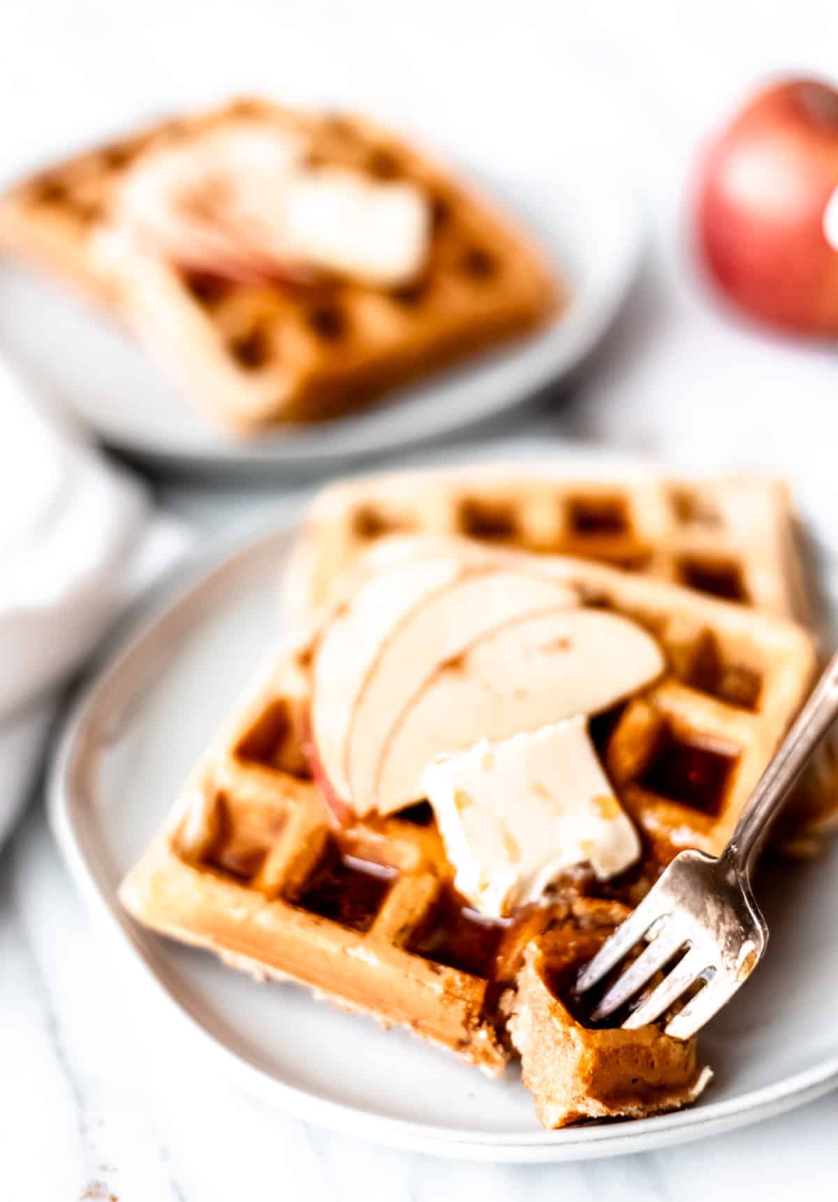 A fork with a bit of apple cinnamon waffle on it in front of 2 waffles stacked on a plate and topped with butter, syrup and apple slices.