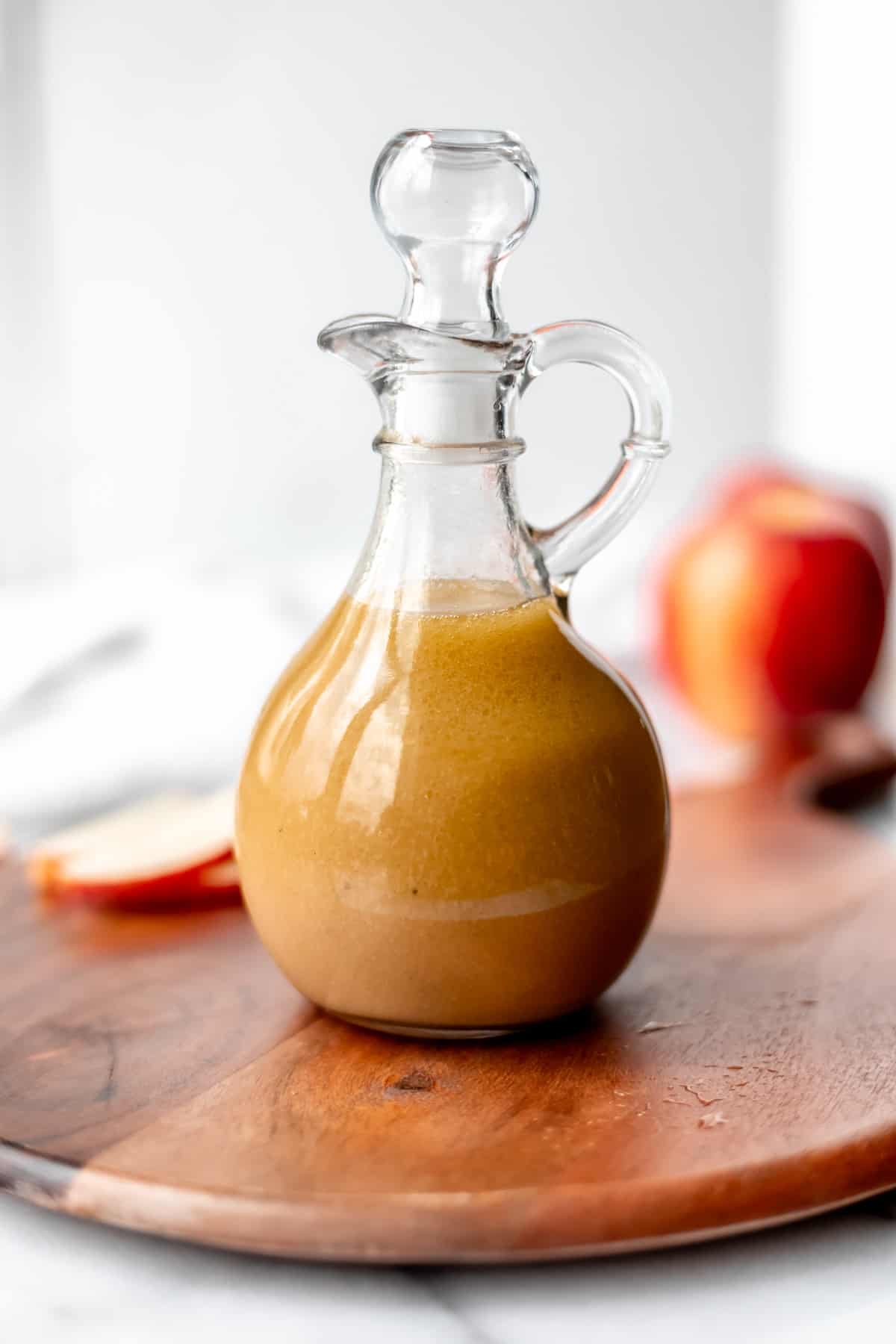 Apple maple vinaigrette in a cruet with a lid on it on a wood board with an apple and apple slices in the background.