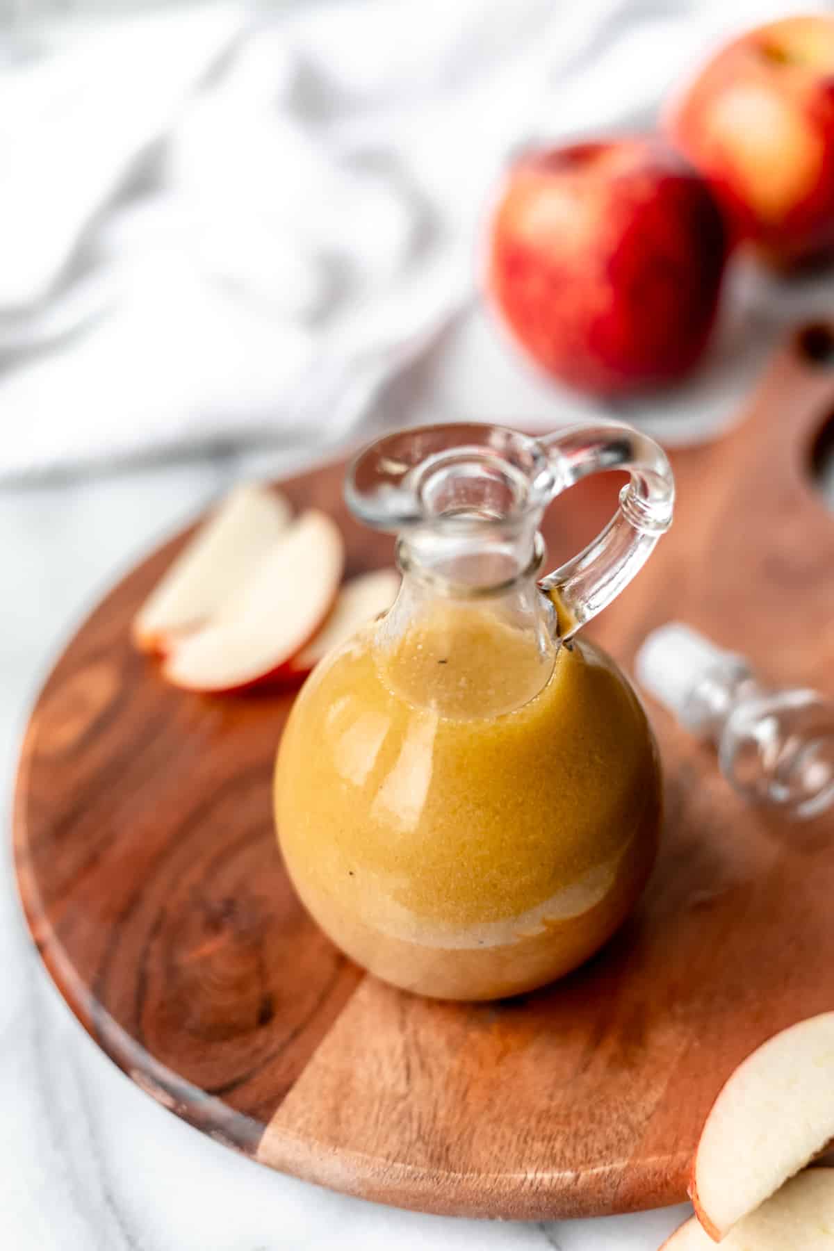 Apple maple vinaigrette in a cruet with the lid off next to it on a wood board with an apple and apple slices in the background.