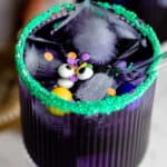 Close up of a blue curacao Halloween cocktail garnished with sprinkles and candy eyes with text overlay.