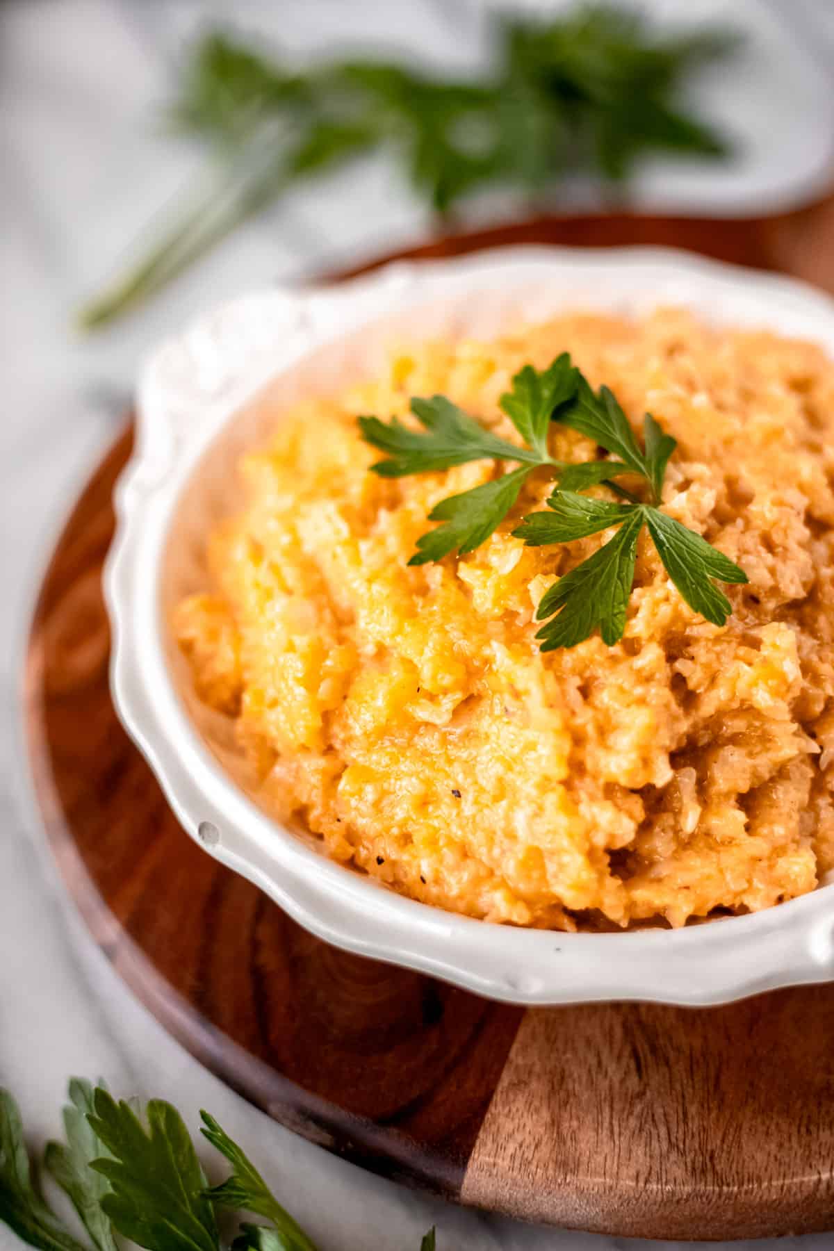 Close up of cheesy cauliflower rice in a white bowl on a wood server with fresh parsley around it.